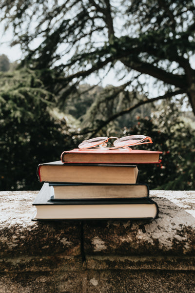 a stack of books with a pair of glasses on top. Sitting on a stone wall with trees in the background.